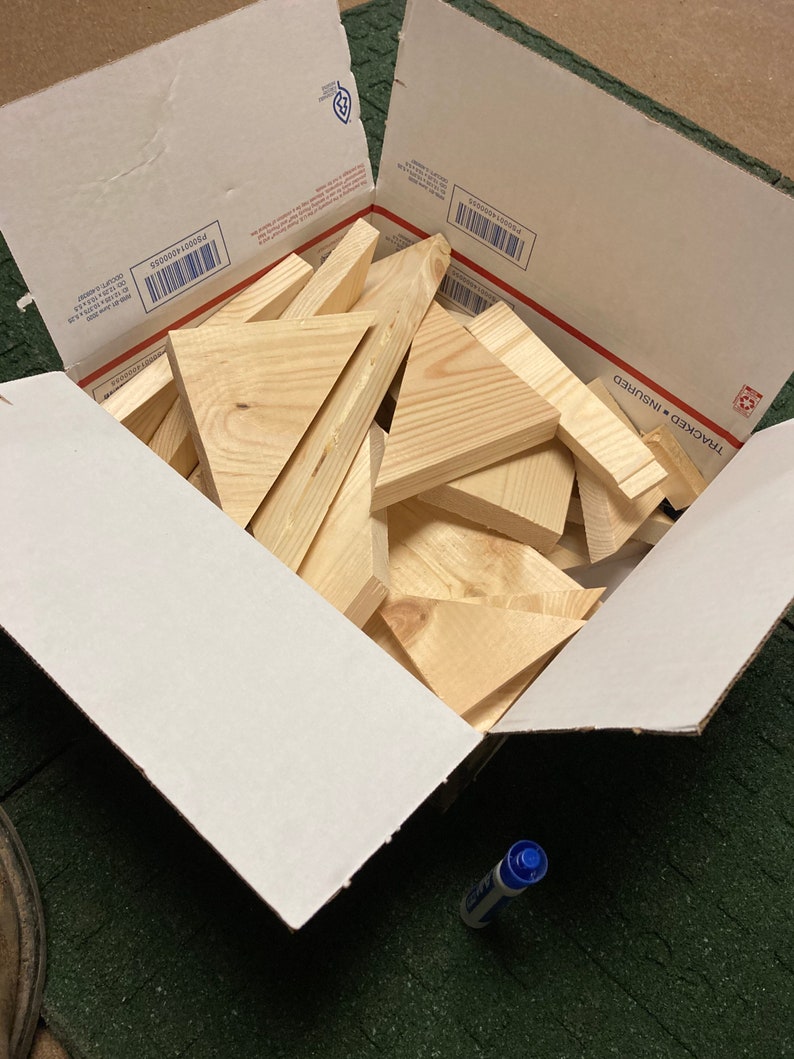 May include: A cardboard box filled with a variety of wooden triangles. The box is open and the triangles are visible inside. The box is sitting on a green surface.