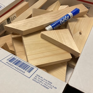 May include: A box filled with various sizes of unfinished wooden triangles and rectangles. The box is labeled with the text "RRB-B1 June 2020", "ID: 12.125 x 10.375 x 5.25", "OD: 12.25 x 10.5 x 5.5", "ODCUFT: 0.409397", and "PS00014000055". A blue dry-erase marker is lying on top of the wood pieces.