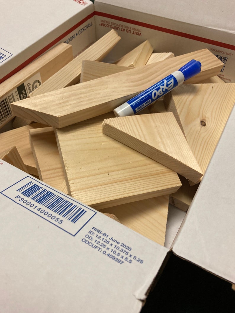 May include: A box filled with various sizes of unfinished wooden triangles and rectangles. The box is labeled with the text "RRB-B1 June 2020", "ID: 12.125 x 10.375 x 5.25", "OD: 12.25 x 10.5 x 5.5", "ODCUFT: 0.409397", and "PS00014000055". A blue dry-erase marker is lying on top of the wood pieces.