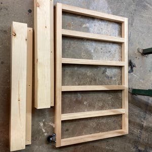 K&ouml;nnte beinhalten: Unfertiger Holzrahmen mit horizontalen Lamellen und zwei vertikalen St&uuml;tzst&uuml;cken sowie zwei weiteren Holzst&uuml;cken. Der Rahmen hat unten ein Rad. Das Holz hat eine nat&uuml;rliche, helle Farbe.