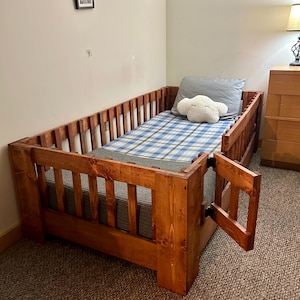 Puede incluir: Una cama de madera para niños pequeños con una cabecera y un piecero de listones. La cama tiene un colchón a cuadros azules y blancos y una almohada gris con un diseño de nube blanca. La cama tiene una puerta de seguridad en un lado que se puede abrir y cerrar.