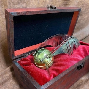 Op de afbeelding: Een gouden replica van de Snitch met zilveren vleugels rust op rood fluweel in een donkerbruine houten kist. De kist heeft een rode binnenrand en een metalen sluiting.