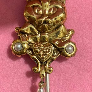 Puede incluir: Un broche de tono dorado con la cabeza de un gato con un corazón y detalles de perlas. El gato lleva una cinta alrededor del cuello.