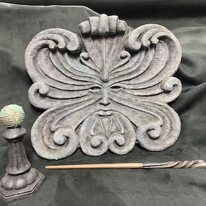 May include: A wooden wall plaque featuring a carved green man face. The plaque is adorned with intricate details and has a weathered finish. A small, round, green and brown orb sits on a black pedestal in front of the plaque.