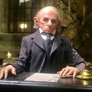 Op de afbeelding: Een man met grote oren en een gerimpeld gezicht draagt een donkergrijs pinstripe pak met een wit overhemd en een donkere das. Hij zit aan een bureau met een vel papier voor zich. Er staat een gouden inktpot en een pen op het bureau.