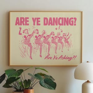 May include: A framed print with the text "ARE YE DANCING?" in pink, above a line of dancing women in pink outfits. The print is in a wooden frame and hangs above a wooden cabinet with plants and a lamp.