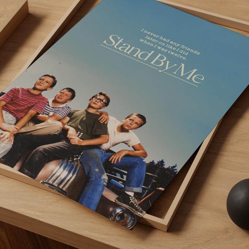 May include: A poster for the movie "Stand By Me" featuring a group of young boys. The poster has a blue sky background and the movie title in large white letters. The quote "I never had any friends later on like I did when I was twelve" is also visible.