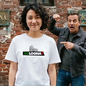 Può includere: Maglietta bianca con un'illustrazione in bianco e nero del Colosseo e la scritta "BOLOGNA?" in verde, nero e rosso. La persona indossa la maglietta davanti a un muro di mattoni. Un'altra persona indica.