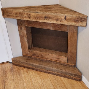 Pu&ograve; includere: Un camino a angolo in legno rustico con un'apertura rettangolare per un inserto per camino. Il camino &egrave; realizzato in legno riciclato con una finitura naturale e ha uno scaffale per esporre decorazioni.