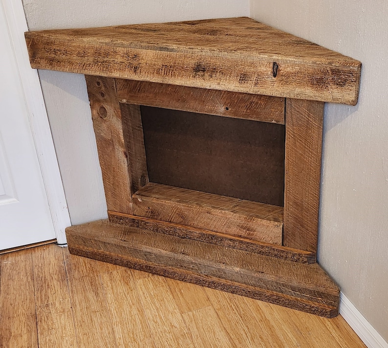 Pu&ograve; includere: Un camino a angolo in legno rustico con un'apertura rettangolare per un inserto per camino. Il camino &egrave; realizzato in legno riciclato con una finitura naturale e ha uno scaffale per esporre decorazioni.