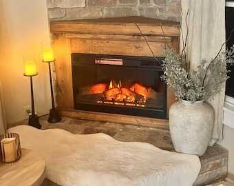 May include: A corner fireplace with a faux fire and a white faux fur rug in front of it. There are two candle holders with lit candles on the left side of the fireplace and a vase with dried branches on the right side.
