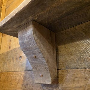 Peut inclure: Gros plan sur une étagère en bois rustique avec un support incurvé. L'étagère et le support sont en bois vieilli, mettant en valeur le grain et la texture naturels. Le support est fixé avec deux goupilles métalliques, ajoutant une touche de design industriel. L'étagère est fixée sur un mur en bois.
