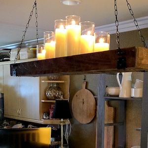 May include: A rustic candle chandelier with a dark wooden beam and hanging chains. The chandelier holds several large, lit candles in glass cylinders, casting a warm, inviting glow. The candles are various heights.