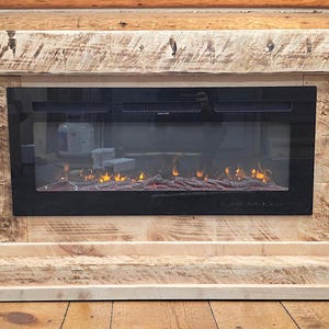 May include: A rectangular electric fireplace with a light-colored, textured natural wood mantel and base. The center features a black glass panel displaying a digital flame effect. Text on the right side reads: Shown With No Stain.