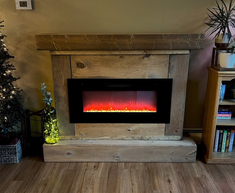 May include: A rectangular electric fireplace insert with a black frame and a simulated glowing orange flame effect, encased in a rustic, light brown wooden mantel surround featuring a thick top shelf and a solid base with visible wood grain.