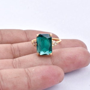 May include: A gold ring with a large, emerald-cut, teal-green gemstone. The ring has smaller, clear gemstones either side of the main stone. The ring is set on a gold band.