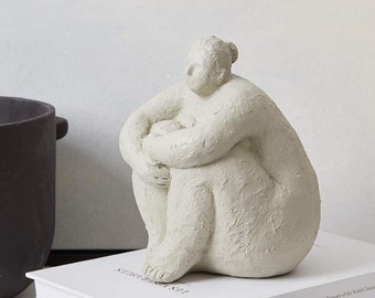 May include: A white ceramic sculpture of a person sitting with their legs crossed and arms wrapped around their knees. The sculpture is sitting on top of two books with the title "Les Miserables" printed on the cover.