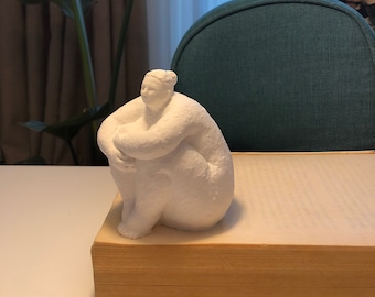 May include: White ceramic sculpture of a person sitting with their arms wrapped around their legs. The sculpture is on a book with a white cover.