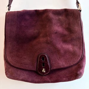 May include: A burgundy suede shoulder bag with a flap closure. The bag features a long, adjustable strap and a decorative stitched pattern on the flap. The bag has a leather accent on the front with a silver-tone clasp.