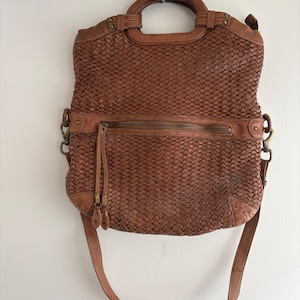 May include: A brown woven leather handbag with a top handle and a detachable shoulder strap. The bag features a zippered pocket across the front and metal hardware. The bag is approximately 12 inches wide and 10 inches tall.
