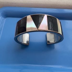 May include: A silver cuff bracelet with a geometric design. The bracelet features alternating black and iridescent white sections, with a polished silver band. The design includes black rectangles and white triangles. The bracelet is open at the bottom.