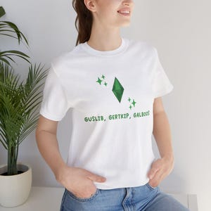 May include: White t-shirt with a green diamond and stars printed on the front. The text "GUSLIB, GERTKIP, GALBOOS" is printed below the diamond.