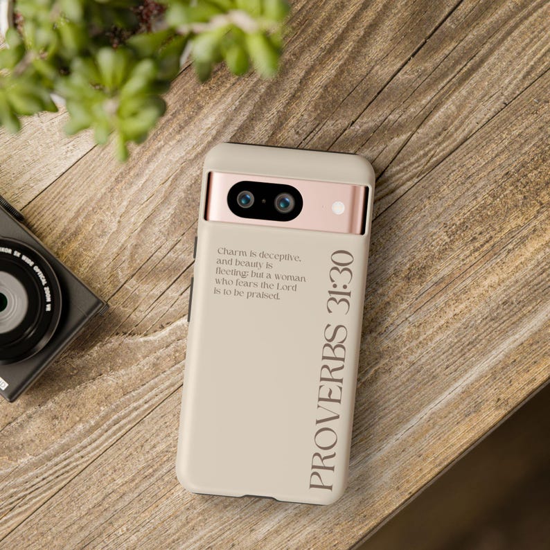 Puede incluir: Una funda de smartphone beige con una barra de c&aacute;mara en oro rosa. La funda presenta el texto 'Charm is deceptive, and beauty is fleeting; but a woman who fears the Lord is to be praised.' y el texto vertical grande 'PROVERBS 31:30' en fuente oscura.