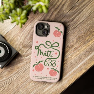 May include: A pink phone case with a design featuring peaches, hearts, and the text "Matt 6:33." The case has a green bow and the quote "But seek first his kingdom and his righteousness, and all these things will be given to you as well."