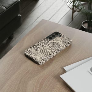 May include: A phone case with a beige and black pattern featuring text from John 3:16. The case has a black camera cutout and is lying on a light wood surface. The design includes floral and zebra stripe elements.