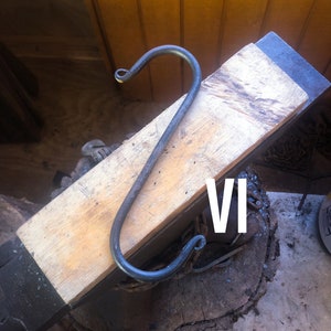 May include: A metal hook is being shaped on an anvil. The hook is made of black metal and is bent into a 'S' shape. The anvil is made of wood and has a black metal top. The hook is resting on the anvil with the text 'VI' visible in the background.