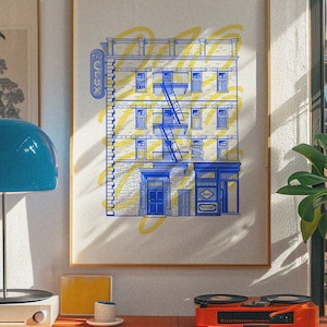 May include: A framed art print depicting a building facade in blue and yellow. The artwork includes a fire escape, windows, and the text "The Cure" on a sign. The print is displayed on a wall above a red shelf.