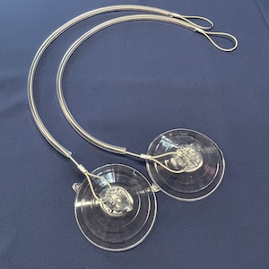 Puede incluir: Dos ventosas transparentes con ganchos de metal plateado unidos a un alambre de metal plateado. El alambre está en forma de círculo.