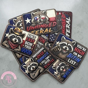 Op de afbeelding: Een verzameling rechthoekige patches met illustraties van wasberen en diverse slogans zoals Unhinged & Feral, Ain't Easy Bein' This Good, Professional Overthinker en You're High On My Bitin List, uitgevoerd in rood, wit en blauw.