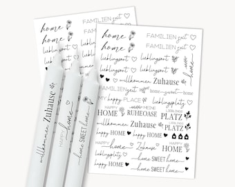 Op de afbeelding: Drie witte kaarsen met zwarte tekst, waaronder "Willkommen Zuhause", "Happy Home" en "Home Sweet Home". Twee vellen stickers met huiselijke zinnen en bloemmotieven zijn ook zichtbaar.