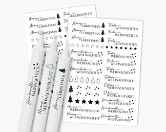 Puede incluir: Tres velas blancas con texto negro "Wundervolle Weihnachten", "Kleines Weihnachtslicht" y "Merry Christmas". Dos hojas de pegatinas con temática navideña están en el fondo.