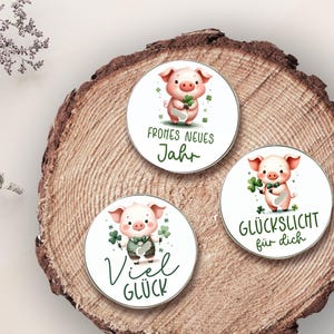 Puede incluir: Tres pegatinas redondas con ilustraciones de acuarela de un cerdo sosteniendo un tr&eacute;bol de cuatro hojas. Las pegatinas tienen texto en alem&aacute;n que dice "Frohes Neues Jahr", "Viel Gl&uuml;ck" y "Gl&uuml;ckslicht f&uuml;r dich". Las pegatinas est&aacute;n sobre una superficie de madera.