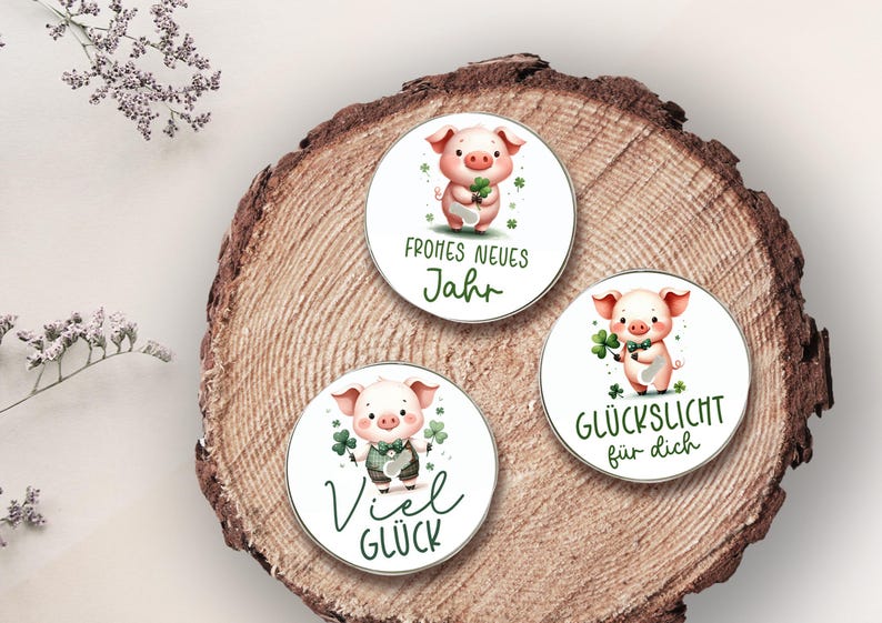 Puede incluir: Tres pegatinas redondas con ilustraciones de acuarela de un cerdo sosteniendo un tr&eacute;bol de cuatro hojas. Las pegatinas tienen texto en alem&aacute;n que dice "Frohes Neues Jahr", "Viel Gl&uuml;ck" y "Gl&uuml;ckslicht f&uuml;r dich". Las pegatinas est&aacute;n sobre una superficie de madera.