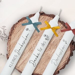May include: Three white candles with black text and floral designs. The candles are tied with blue, yellow, and red ribbons. The text on the candles reads "Kleines Dankesch&ouml;n", "Danke f&uuml;r alles", and "Dankesch&ouml;n".