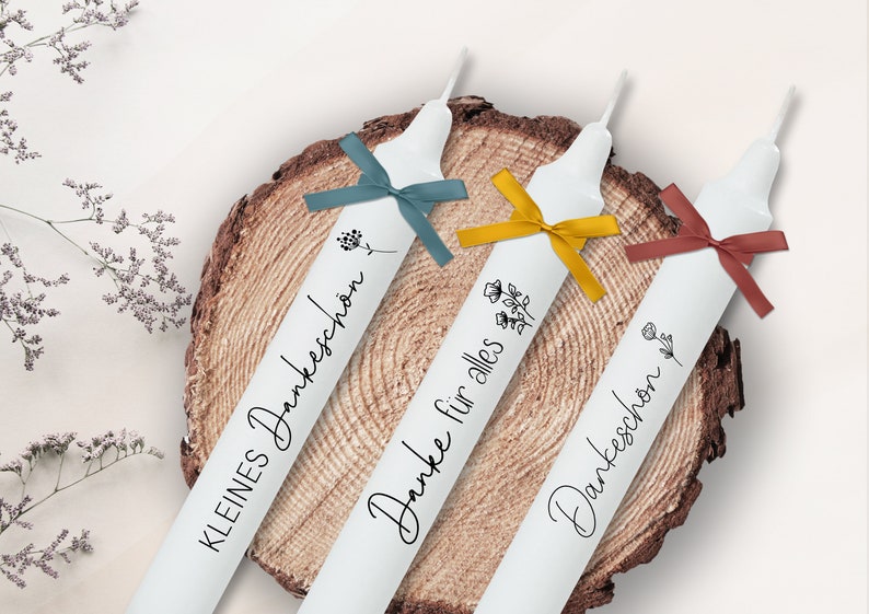 May include: Three white candles with black text and floral designs. The candles are tied with blue, yellow, and red ribbons. The text on the candles reads "Kleines Dankesch&ouml;n", "Danke f&uuml;r alles", and "Dankesch&ouml;n".