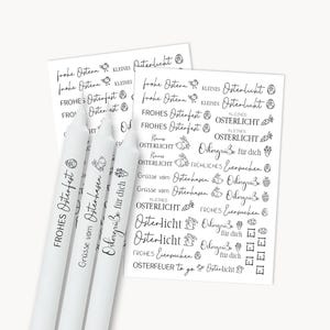 Könnte beinhalten: Drei weiße Stabkerzen mit schwarzer Schrift, neben zwei Papierbögen mit Ostermotiven und Illustrationen. Die Kerzen tragen die Aufschrift "Frohes Osterfest". Das Papier enthält Sätze wie "Frohe Ostern" und "Osterlicht".