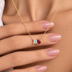 May include: A delicate gold chain necklace featuring a pendant with two stones: a clear square-cut gemstone and a vibrant red pear-shaped gemstone. The pendant is set in a gold-toned metal frame, suspended from a thin, polished gold link chain.