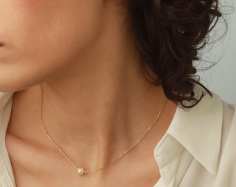 May include: A delicate gold chain necklace featuring a single small white pearl pendant, paired with a matching white pearl stud earring. The jewelry set is worn against a plain white collared shirt, highlighting the simple and refined design.