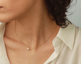 May include: A delicate gold chain necklace featuring a single small white pearl pendant, paired with matching white pearl stud earrings. The necklace rests against the neck, while the earrings are worn in the earlobes.