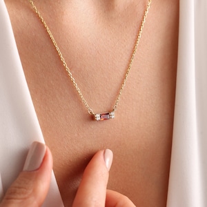 May include: A gold chain necklace featuring a horizontal bar pendant. The pendant is set with a central rectangular baguette-cut red gemstone flanked by two small clear stones on each side, all held within a polished gold metal setting.
