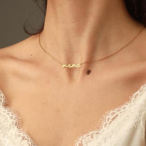 May include: A gold-toned necklace featuring a delicate chain and a central pendant in cursive script that spells out the word mama. The necklace rests against the neck, showcasing a polished metallic finish and a simple, refined design.