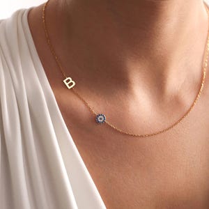 May include: A gold necklace with the letter "B" and a blue and white evil eye charm. The fine chain is worn around the neck, displaying the pendant's design. The necklace is a piece of jewellery.