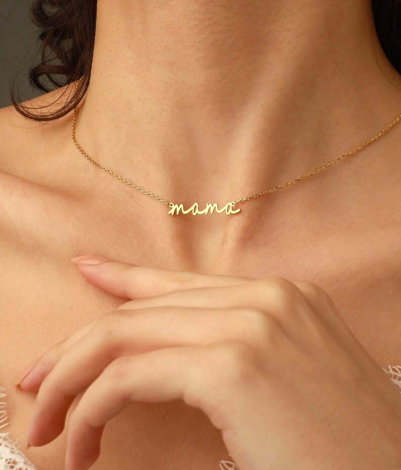 May include: A gold-toned necklace featuring a delicate chain and a central pendant in a cursive script font that spells out the word mama. The metal has a polished finish, and the pendant is integrated directly into the thin, lightweight chain design.