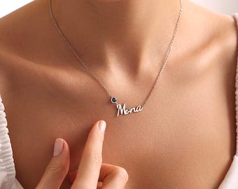 May include: A silver-toned necklace featuring a delicate chain and a script-style nameplate pendant that reads Mona. The necklace includes a small, circular, dark blue gemstone accent positioned directly next to the nameplate on the chain.