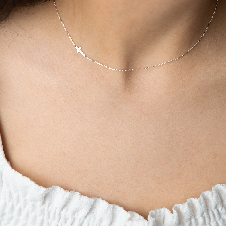 K&ouml;nnte beinhalten: Eine zarte silberne Halskette mit einem kleinen Kreuzanh&auml;nger, der seitlich positioniert ist. Die Halskette wird vor einem hellen Hintergrund pr&auml;sentiert, wodurch die feine Kette und das schlichte Design des Kreuzes hervorgehoben werden. Ein subtiles und elegantes Schmuckst&uuml;ck.