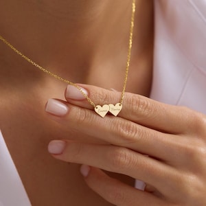 Könnte beinhalten: Eine goldfarbene Halskette mit zwei herzförmigen Anhängern. Jeder Anhänger trägt die Namen "Emma" und "Henry". Die Kette wird auf der Brust und der Hand einer Person gezeigt.
